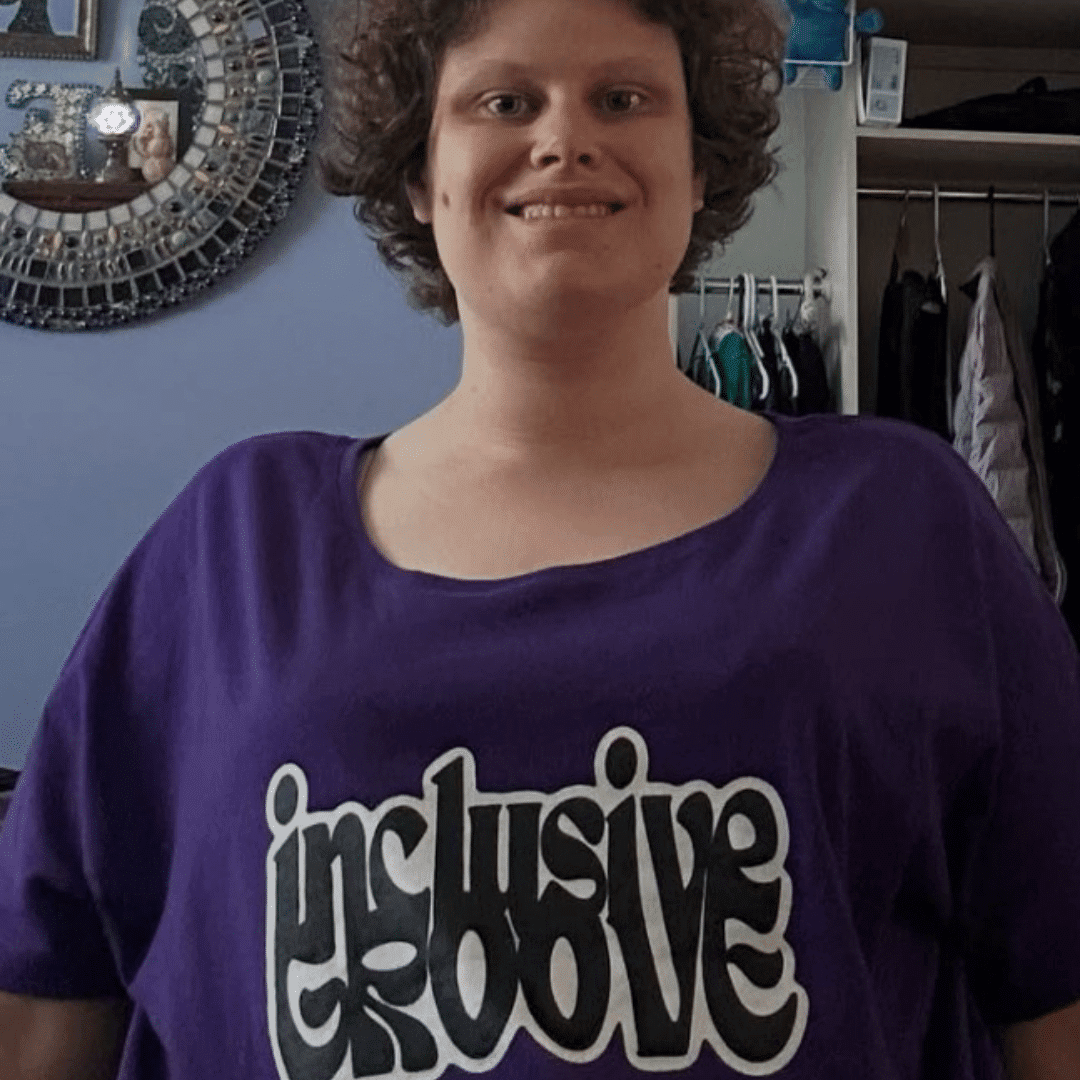 A woman in her late 20s wearing a T-shirt with the words Inclusive Groove smiles at the camera.