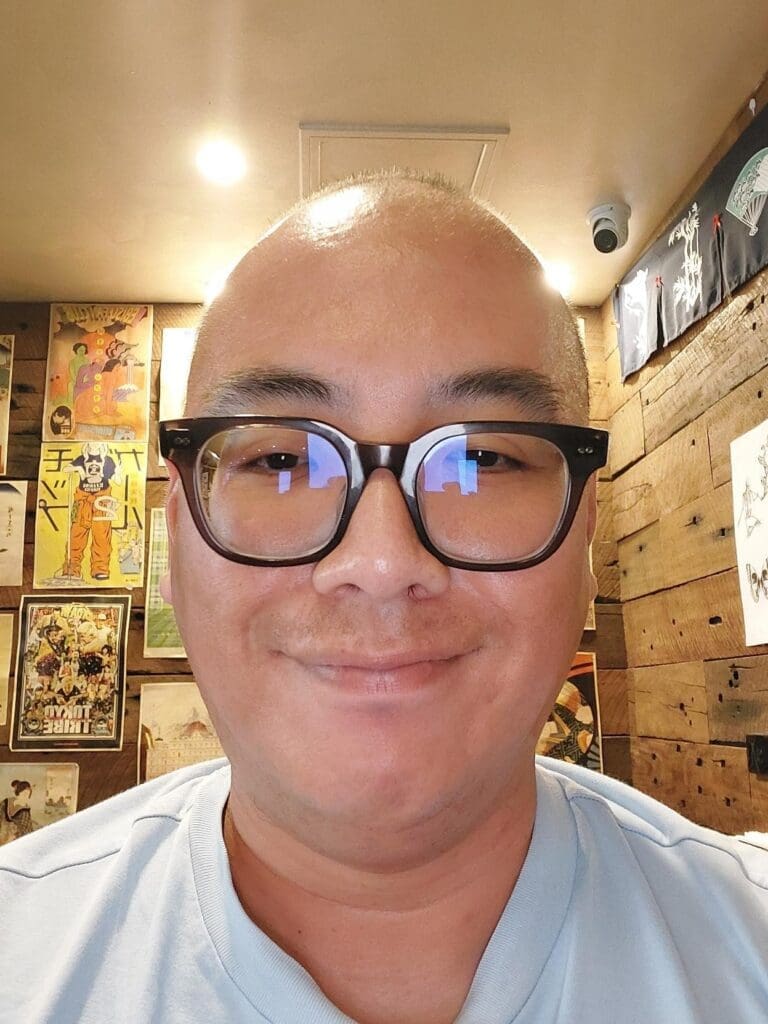 A young man smiles broadly at the camera. He is wearing glasses with dark cat eye frames and a light blue T-shirt.