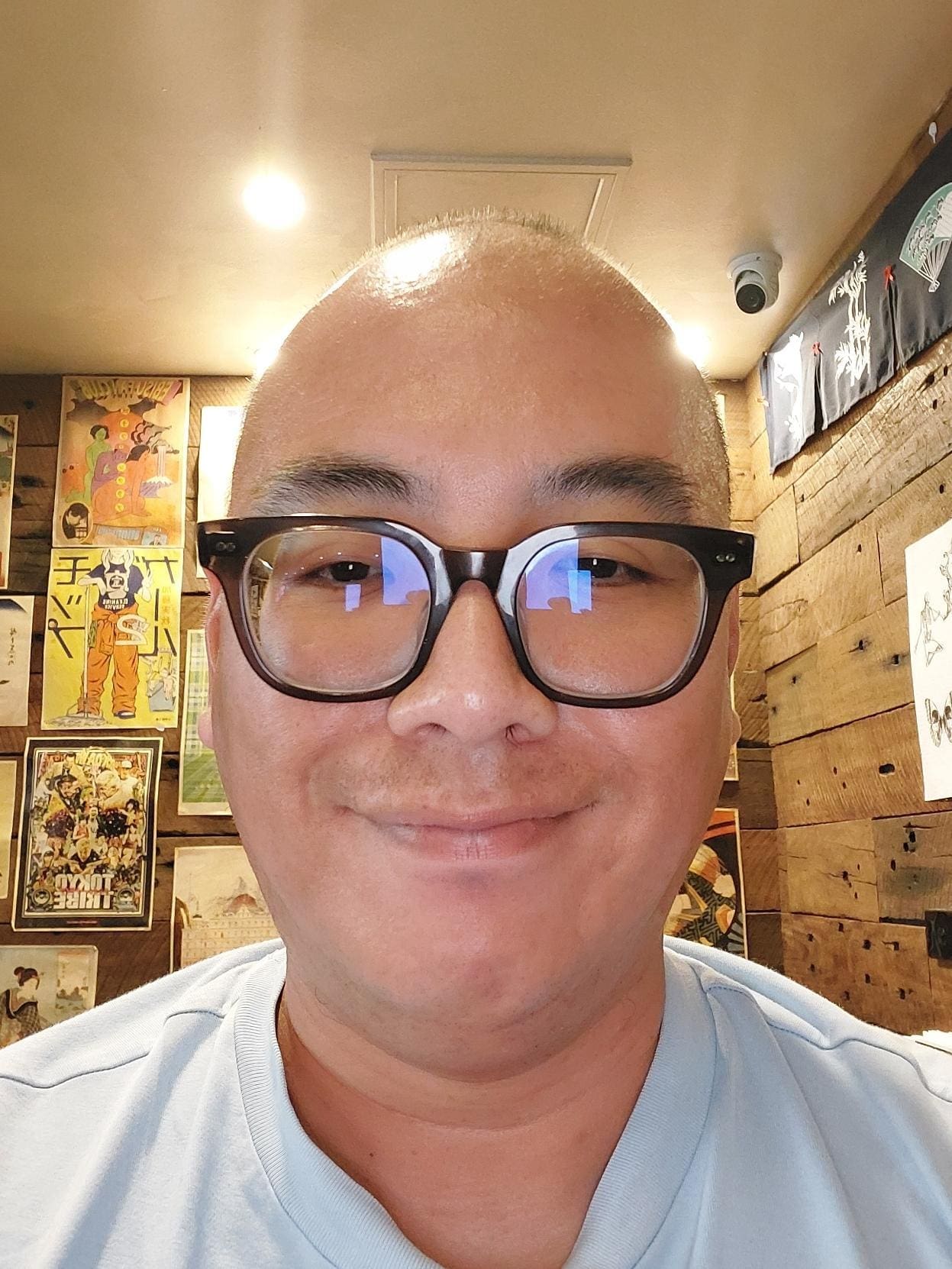 A young man smiles broadly at the camera. He is wearing glasses with dark cat eye frames and a light blue T-shirt.