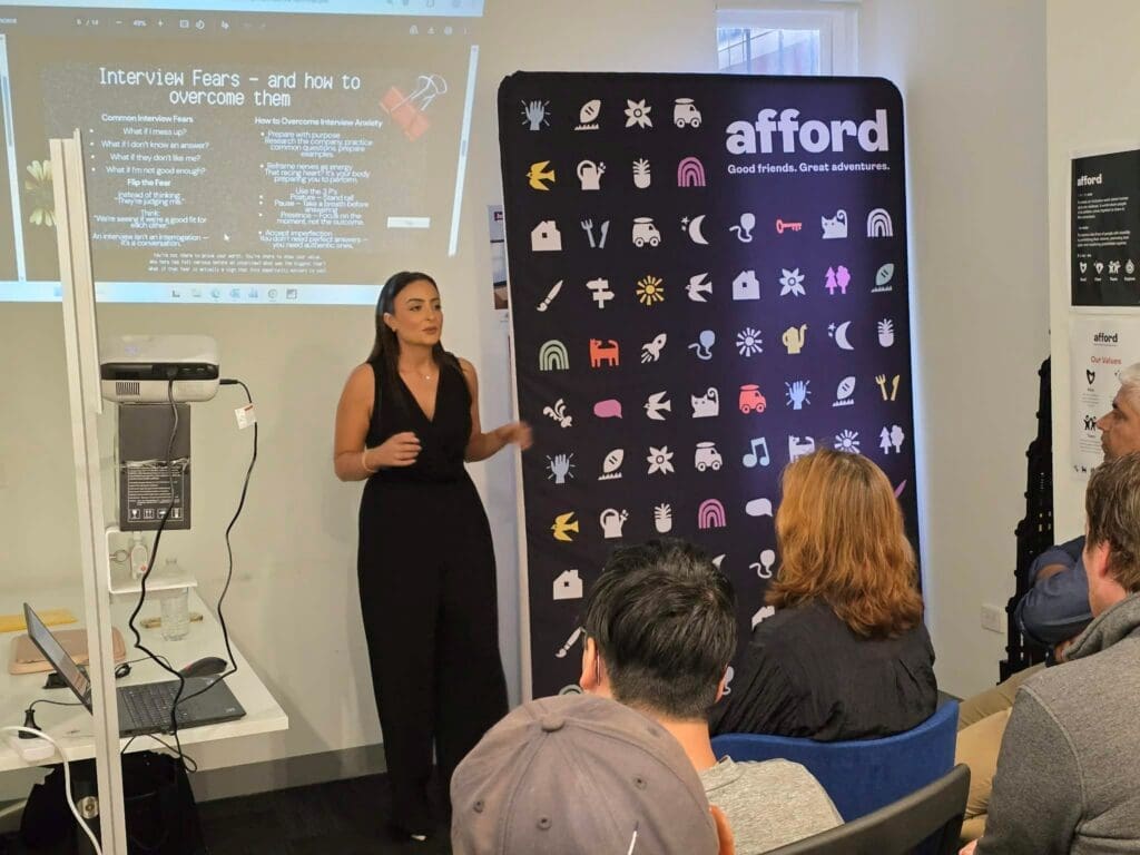 A woman stands in front of a group of people who are seated and listening intently. She is talking and presenting on a whiteboard.