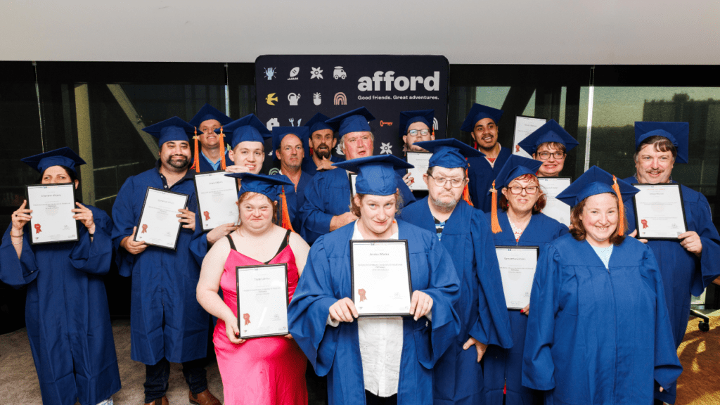 A group of people with disability wear royal blue graduation caps and gowns and hold certificates while smiling directly at the camera. 