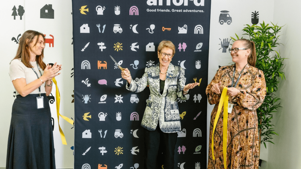 A woman holds scissors up high as a yellow ribbon drops to the floor. A second and third woman stand to each side and clap with warm applause. 