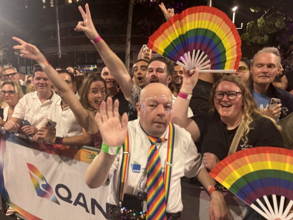 A man smiles at the camera waving his hand hello. He is wearing a rainbow coloured tie and behind him is a crowd of fans many waving rainbow flags and fans.