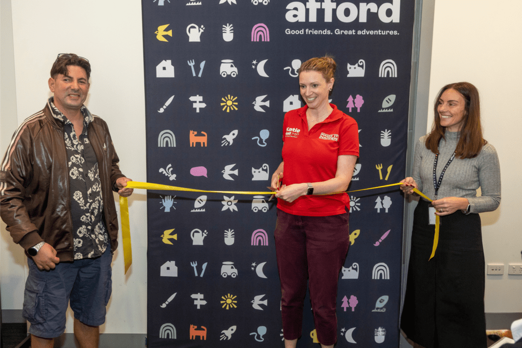 A man stands holding a ceremonial ribbon with two women. The woman in the centre is holding gold scissors and is about to cut the ribbon. There's an Afford banner behind them. There's a celebratory vibe.