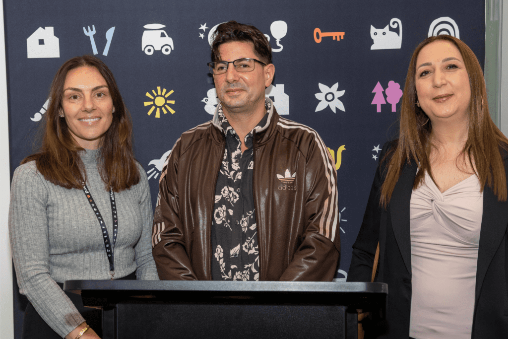 Two women stand on either side of a man. All three are smiling broadly at the camera and look very happy. The man in the centre appears prooud of himself.