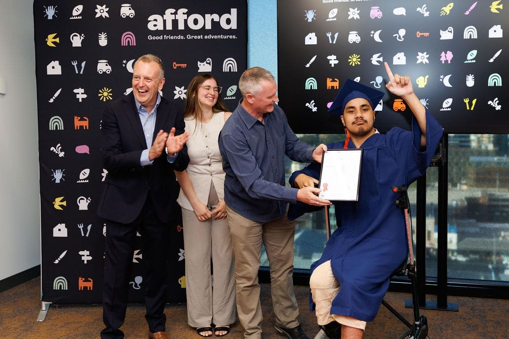 A young man wearing a graduation cap and gown holds his certificate and one finger in up in celebration. The mood is joyous. Behind him is a banner saying Afford in bright white letters.