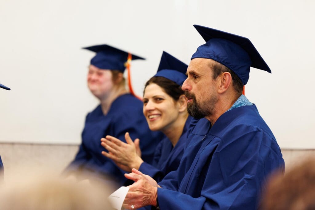 A woman in a blue graduation cap and gown smiles warmly while clapping her fellow students.